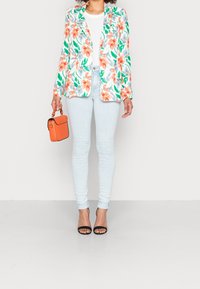 Żakiet w kwiatowy wzór w odcieniach pomarańczowym i zielonym, w połączeniu z jasnoniebieskimi rurkami i czarnymi sandałami na obcasie, trzyma pomarańczową torebkę.
