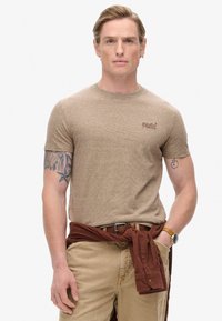 Camiseta marrón de manga corta con un patrón texturizado, cuello redondo y un pequeño logo bordado. Combinada con pantalones beige y una camisa marrón atada en la cintura.