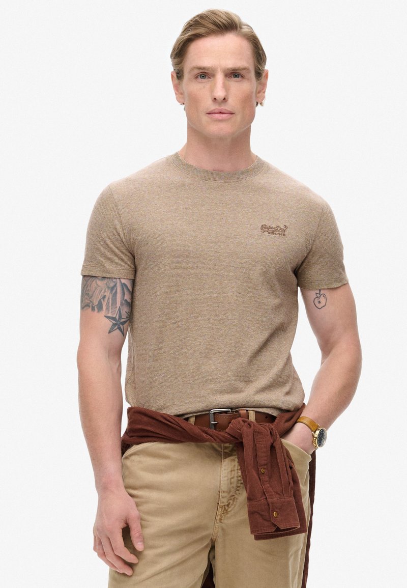 Camiseta marrón de manga corta con un patrón texturizado, cuello redondo y un pequeño logo bordado. Combinada con pantalones beige y una camisa marrón atada en la cintura.