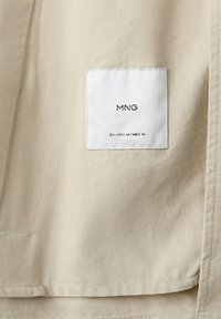 Tessuto beige con un'etichetta quadrata bianca cucita sopra, che mostra "MNG" e i dettagli della taglia "EU-USA M/MEX M" in testo nero.