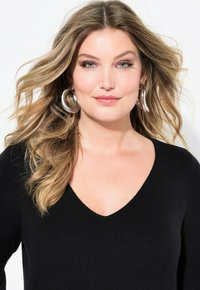 Maglione nero a coste con scollo a V profondo. Il modello ha capelli biondi mossi e grandi orecchini a cerchio argentati e lucidi. Pelle liscia e trucco naturale.