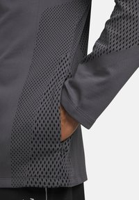 Gray athletic top featuring breathable mesh panels with circular cutouts, and a relaxed fit. Includes a side pocket for storage.