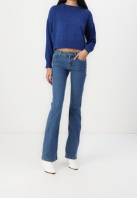 Pull court bleu avec ourlet côtelé, assorti à un jean évasé bleu. Le mannequin porte des bottines blanches à talons. Textures lisses, design décontracté.