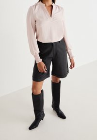 Ljusrosa blus med lång ärm och V-ringning, smockade manschetter, kombinerad med skräddarsydda shorts i mörka ränder och knähöga svarta läderstövlar.