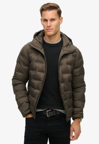 Gevoerde olijfgroene jas met een capuchon, met een ritsluiting en elastische boorden, gedragen over een donker gebreid trui. Zwarte riem en jeans zichtbaar.