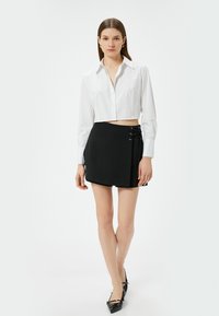 Camicia bianca cropped con bottoni e maniche lunghe, abbinata a una mini gonna nera con dettagli di lacci laterali. Il modello indossa scarpe basse.