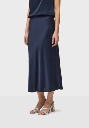 Donna che indossa una gonna midi blu navy e sandali argento con tacco alto, in piedi contro uno sfondo chiaro e semplice.