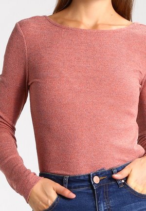 Kvinde iført en tætsiddende, langærmet, ribbet lyserød top, der er stoppet ned i højtaljede blå jeans med hænderne i lommerne.