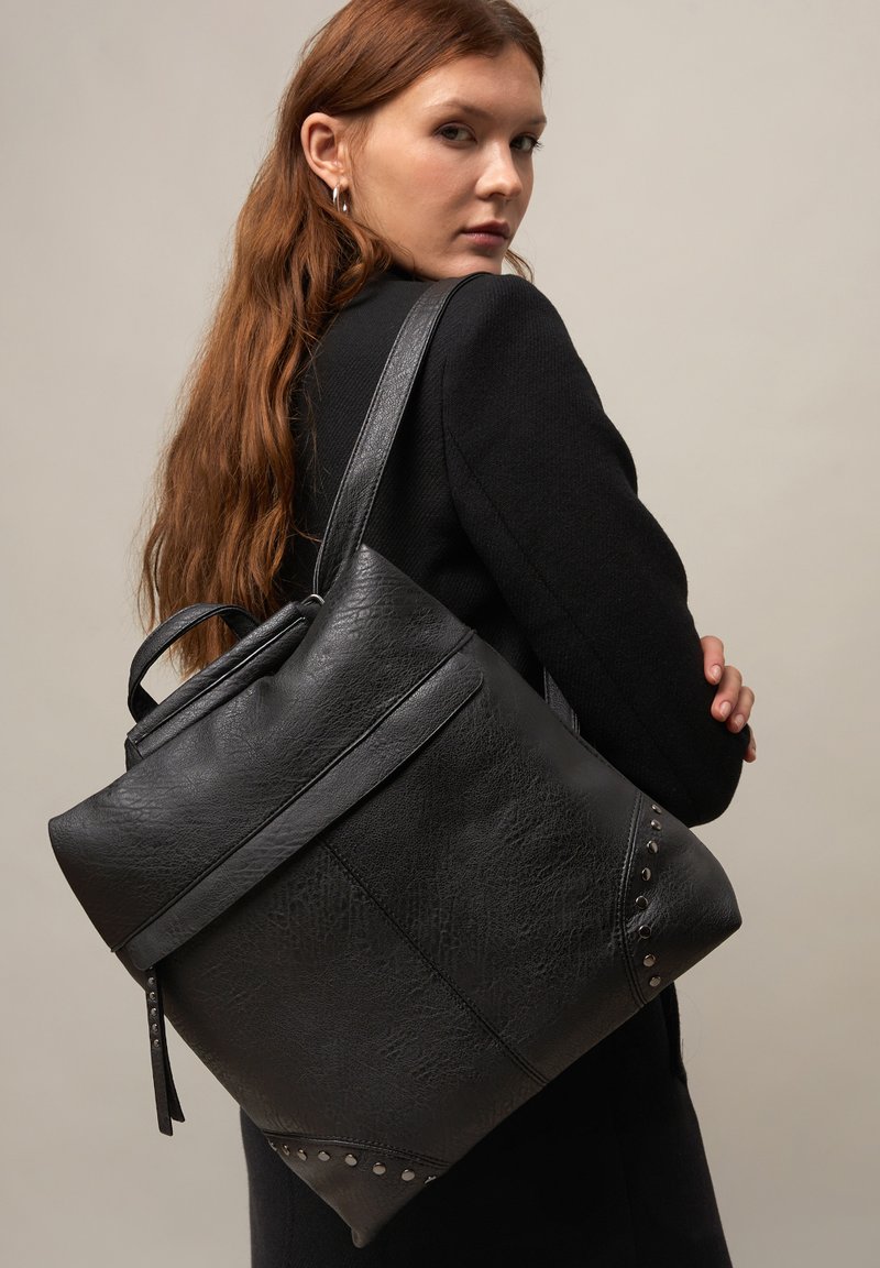 Žena s dlhými červenými vlasmi, oblečená v čiernom, drží veľkú čiernu štruktúrovanú koženú kabelku s cvočkami na rohoch cez rameno.