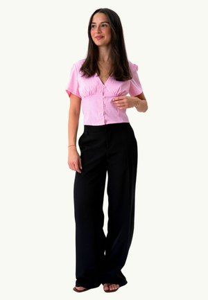 Jeune femme aux longs cheveux foncés portant un chemisier rose à manches courtes et boutonné ainsi qu’un pantalon large noir, debout avec un léger sourire.