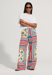 Un pantalon large multicolore présente un motif géométrique vibrant avec des accents rouges, bleus, jaunes et verts ; associé à une chemise blanche simple.