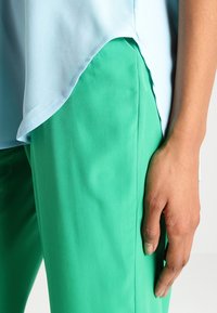 Lyseblå bluse med en buet søm, parret med grønne bukser. Blusen har en glat tekstur, mens bukserne har en blød finish.