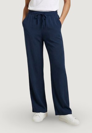 Persona che indossa pantaloni larghi blu navy con vita elasticizzata e coulisse, sneakers bianche e top bianco, con le mani nelle tasche.