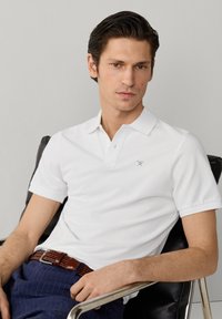 Giovane seduto su una sedia nera indossando una polo bianca con piccolo logo a remi incrociati, pantaloni blu e cintura intrecciata marrone.