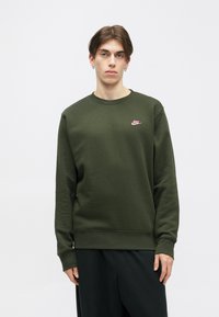 Sweatshirt verde-oliva com gola redonda, mangas longas, punhos canelados e um pequeno logótipo da Nike em rosa no peito. Textura de tecido macio.