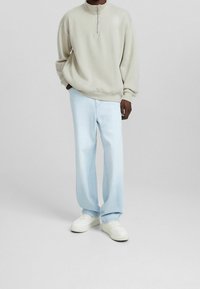 Persona che indossa un felpa beige con zip a un quarto, jeans larghi blu chiaro e sneakers bianche, in piedi davanti a uno sfondo neutro.