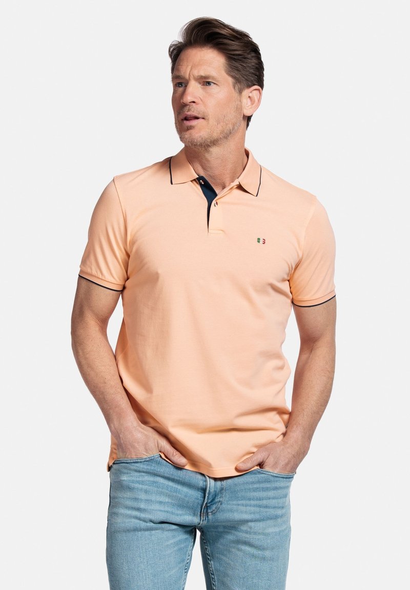 Polo shirt couleur pêche avec un col marine, des manches courtes et un petit logo. Le tissu semble lisse et léger.
