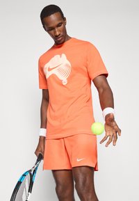 Πορτοκαλί βαμβακερό T-shirt με λευκό λογότυπο Nike. Συνδυασμένο με πορτοκαλί σορτς, κρατώντας μια πράσινη τένις μπάλα με μια ρακέτα.