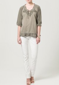 Blusa verde oliva con pizzo e orlo a volant, maniche a tre quarti, abbinata a jeans bianchi attillati e sandali di colore chiaro.