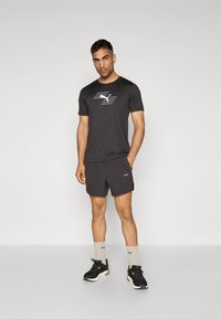 Svart tränings-t-shirt med logga på bröstet, kombinerad med svarta shorts. Bärs med svarta och gröna sportskor samt grå strumpor.