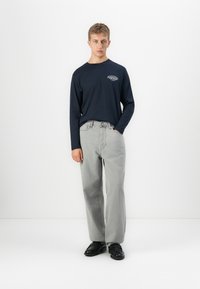 Ung man står framför en vit bakgrund iförd en marinblå långärmad tröja med vitt tryck, ljusgrå jeans och svarta skor, med ena handen i fickan.