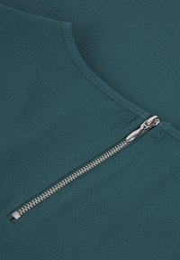Top en tissu bleu sarcelle avec une texture lisse, présentant une fermeture éclair en argent visible sur un côté. Le design a un col rond et un style minimaliste.