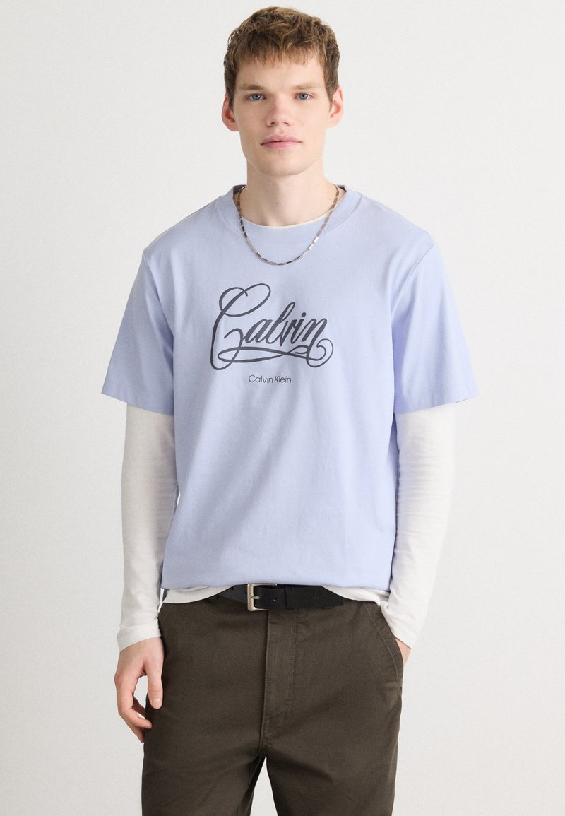 Jasnoniebieska bawełniana koszulka z logo "Calvin Klein" w czarnym skrypcie, nałożona na białą koszulę z długim rękawem, zestawiona z ciemno-brązowymi spodniami.