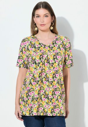 Kvinna iklädd en kortärmad V-ringad blus med blommönster i rosa och gult, kombinerad med mörkblå jeans och prydd med örhängen i form av prästkragar.