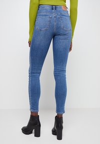 Vaqueros ajustados de mezclilla azul con cintura alta, que presentan dos bolsillos traseros y una etiqueta de cuero marrón. Combinados con botines negros de tacón.