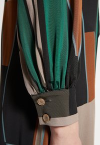 Robe à manches longues présentant un motif géométrique multicolore. Les matériaux comprennent un tissu soyeux avec des manches froncées et des détails de bouton marron.
