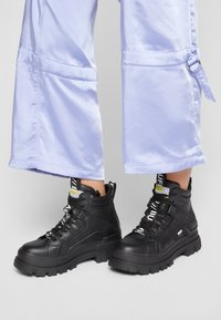 Schwarze Knöchelschäfte aus strukturiertem Leder, mit stabilen Sohlen und kontrastierenden Schnürsenkeln. Kombiniert mit helllila weit geschnittenen Hosen.