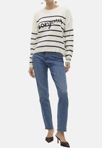 Maglione a righe color crema con linee blu navy e testo, abbinato a jeans azzurri e sandali aperti con decorazioni.