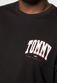 Camiseta de algodón negra con cuello redondo. Presenta un atrevido logo rojo de "TOMMY" y "NEW YORK CITY" en blanco debajo, con un pequeño acento a rayas.