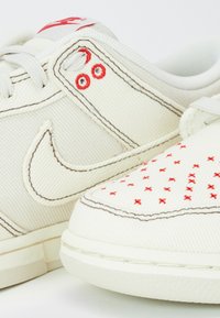 Baskets en toile blanche avec des surpiqûres rouges et des motifs décoratifs brodés en forme de croix rouges, dotées d'une semelle en caoutchouc et d'un tissu texturé.