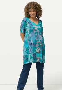Tunika turchese con maniche corte, caratterizzata da motivi astratti vari in blu e bianco; abbinata a jeans blu scuro.