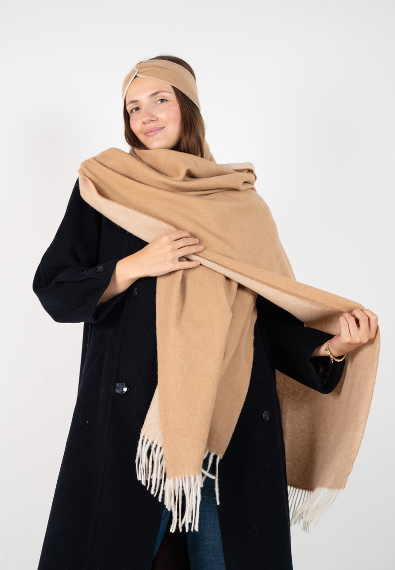 Beige Wollschal mit Fransen, um die Schultern gewickelt, kombiniert mit einem passenden Stirnband, über einem schwarzen Mantel getragen. Glatte Textur und weiches Erscheinungsbild.