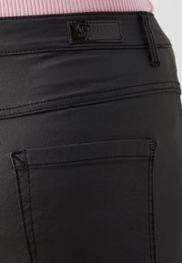 Gros plan sur un pantalon noir en simili cuir avec poche arrière et étiquette de marque, porté avec un haut côtelé rose.