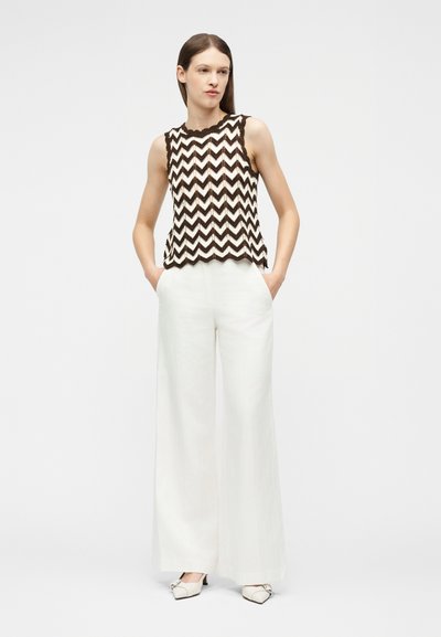 Γυναίκα που φορά τοπ χωρίς μανίκια με ασπρόμαυρο ζιγκ-ζαγκ μοτίβο, λευκό παντελόνι φαρδιάς γραμμής και λευκά παπούτσια με χαμηλό τακούνι, με τα χέρια στις τσέπες, όρθια.