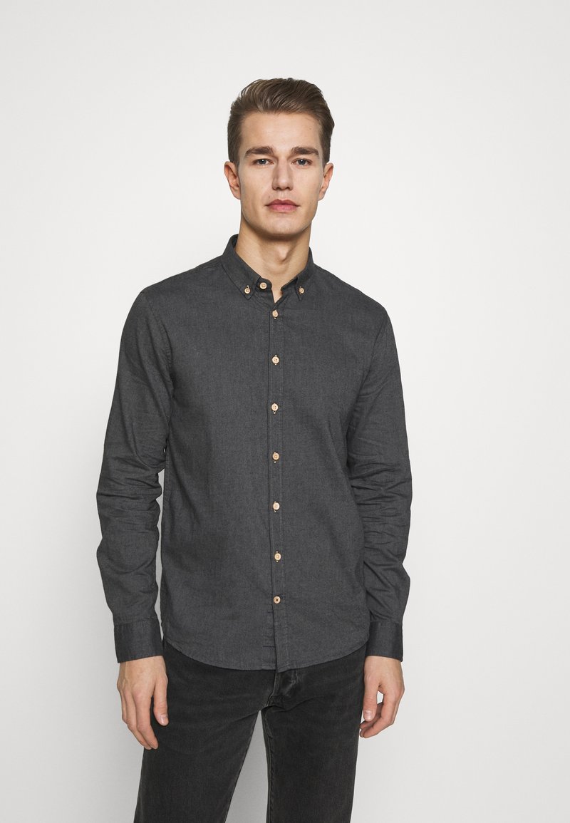 Camisa abotonada de color gris oscuro con cuello, hecha de tela texturizada. Cuenta con botones marrones y mangas largas. Se lleva con unos vaqueros oscuros.