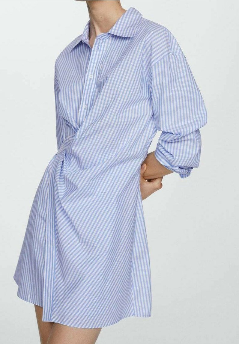 Personne portant une robe-chemise rayée verticale bleu clair et blanc, manches retroussées, mains jointes dans le dos.