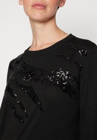 Schwarzer Pullover mit strukturiertem Design und schwarzen Pailletten, die über die Vorderseite verteilt in einem Muster angeordnet sind, Rundhalsausschnitt und lange Ärmel.