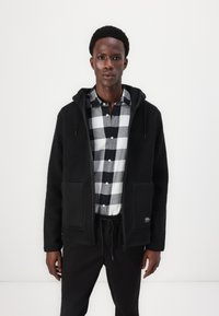 Veste en polaire noire avec capuche, dotée de deux poches avant et d'une bordure en tissu noir lisse. Portée par-dessus une chemise à carreaux noir et blanc.