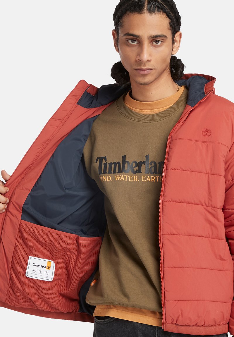 Rote Pufferjacke mit navyblauem Innenfutter, horizontalen Nähten und einer Innentasche. Olivgrüner Sweatshirt mit "Timberland"-Schriftzug.