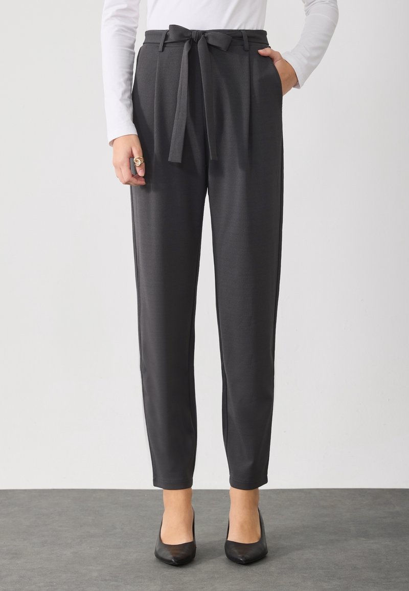 Femme portant un pantalon noir taille haute et fuselé avec ceinture à la taille, un haut blanc à manches longues, des talons noirs et une bague en or à la main gauche.