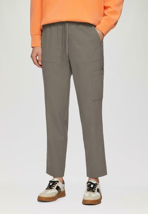 Pantalons jogger gris clair avec une taille élastique, deux poches latérales et une poche cargo sur la cuisse. Associés à des baskets beiges et noires.