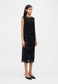 Femme debout portant un haut noir sans manches et une jupe midi en dentelle noire à paillettes, avec des talons noirs à bouts pointus, tournée vers la droite.