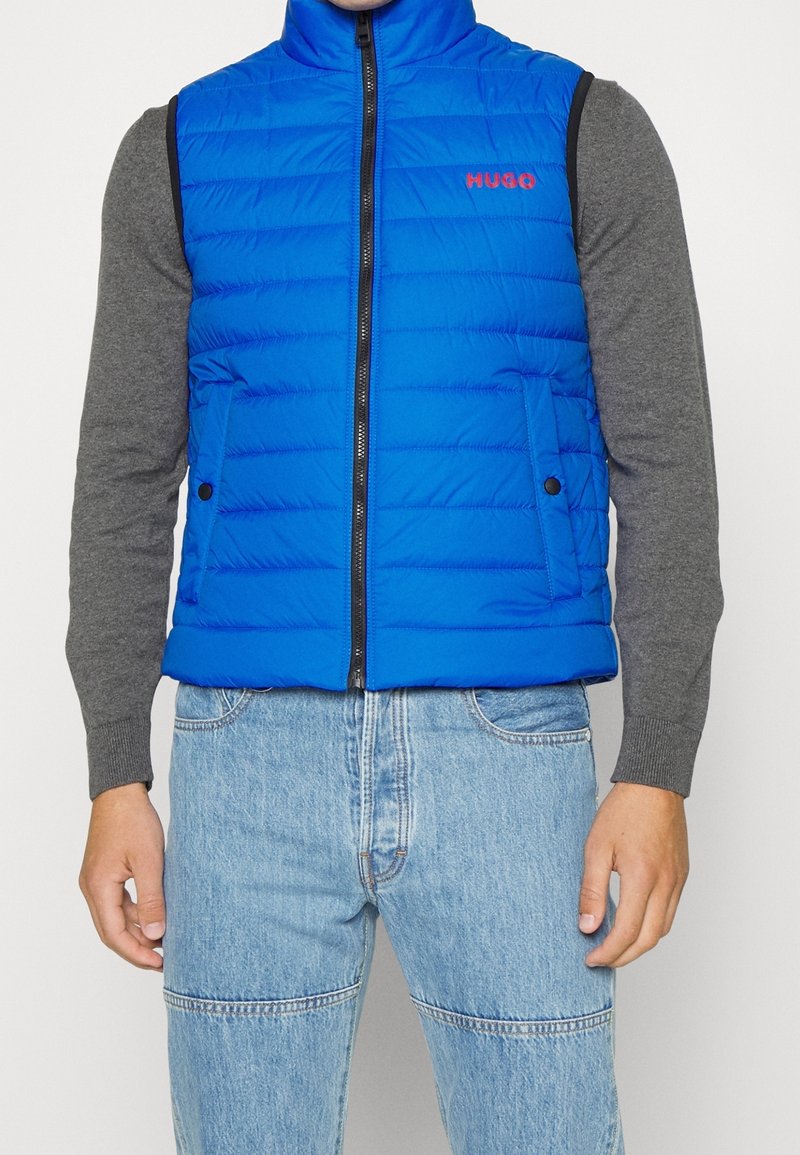 Homme portant une doudoune sans manches bleu vif avec le logo "HUGO", un t-shirt gris à manches longues et un jean denim bleu clair, debout devant un fond uni.