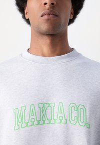 Szürke pulóver bordázott nyakkivágással, nagy zöld nyomtatott "MAKIA CO." felirattal a mellkas területén. Puha, pamut anyag.