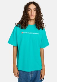 Young person with long curly hair wearing a turquoise shirt with "WIND WATER FIRE EARTH" text and blue denim shorts.