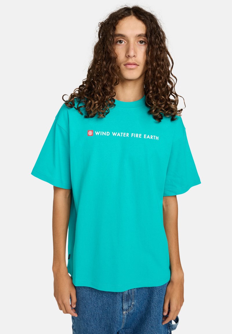 Young person with long curly hair wearing a turquoise shirt with "WIND WATER FIRE EARTH" text and blue denim shorts.